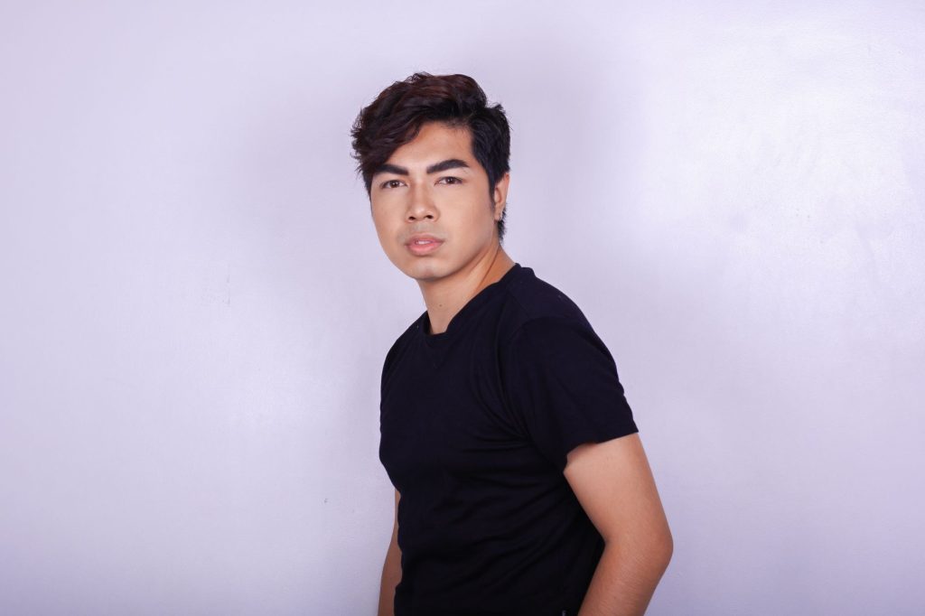 Serious young ethnic man in trendy clothes looking pensively at camera while standing against light lilac wall in modern stud