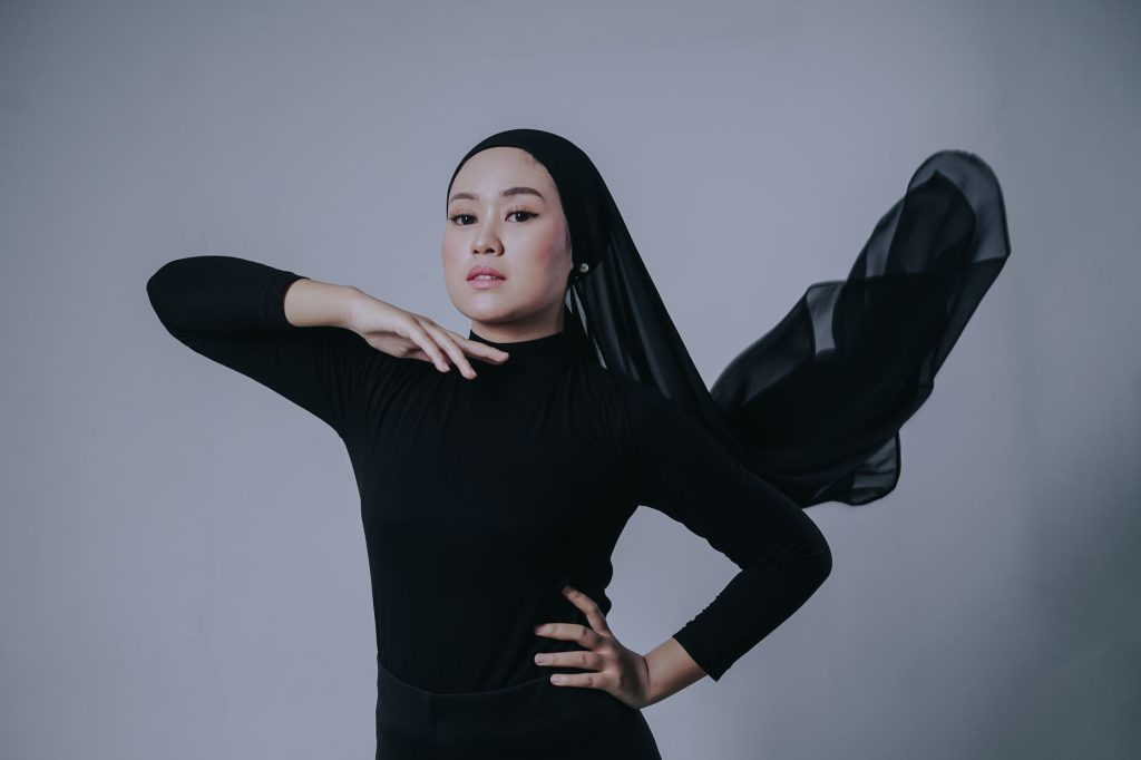 Studio portrait of a confident woman in black with flowing headscarf posing elegantly against a gray background. — Ambiiyah