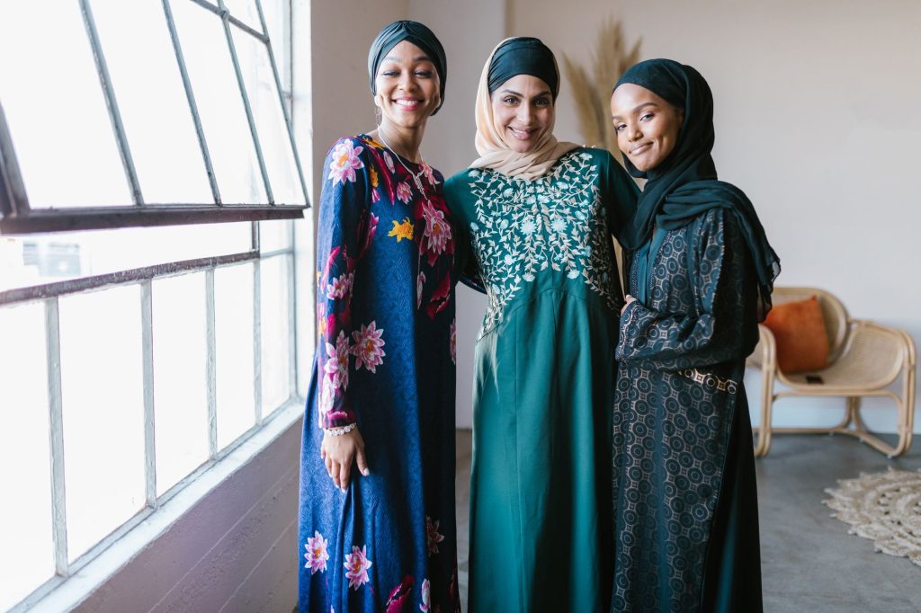 Three women in traditional dresses and hijabs smiling together indoors by a large window. — Ambiyah