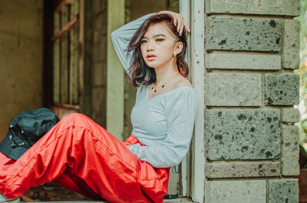 Asian female with hand on head in red pants sitting on window opening in stone wall and looking at camera — Andrea Brillantes