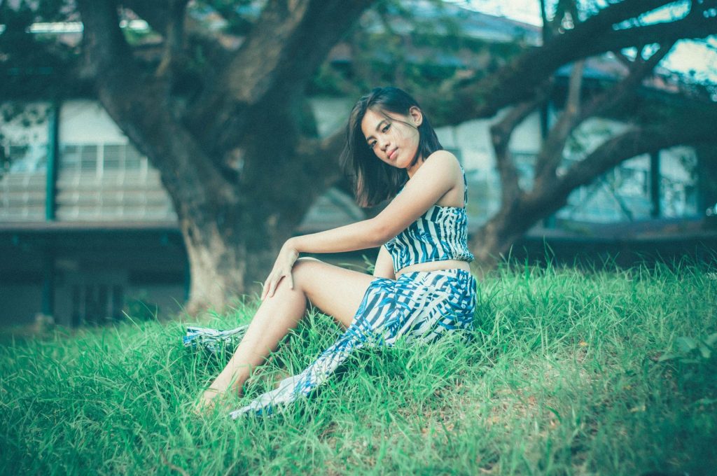 Full body of young Asian female in skirt looking at camera while sitting on grassy ground near tree trunk and house — Asian P