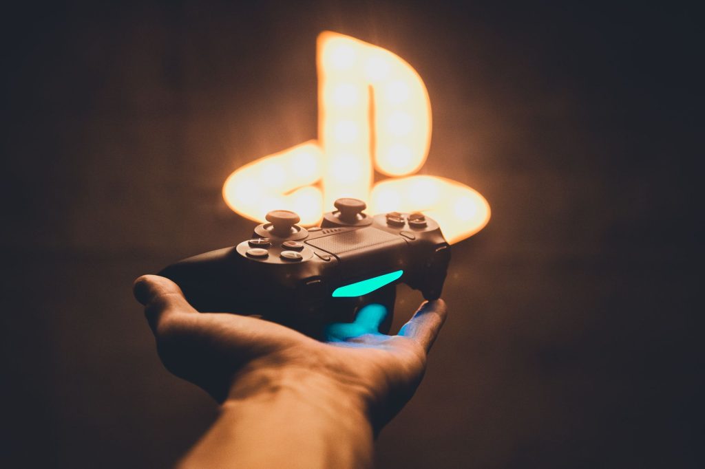 A hand holding a PlayStation controller with a blurred neon game symbol in the background. — Datablitz