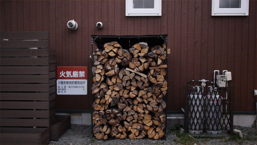 Stack of firewood beside gas cylinders near a brown house wall, no people present. — Itotan