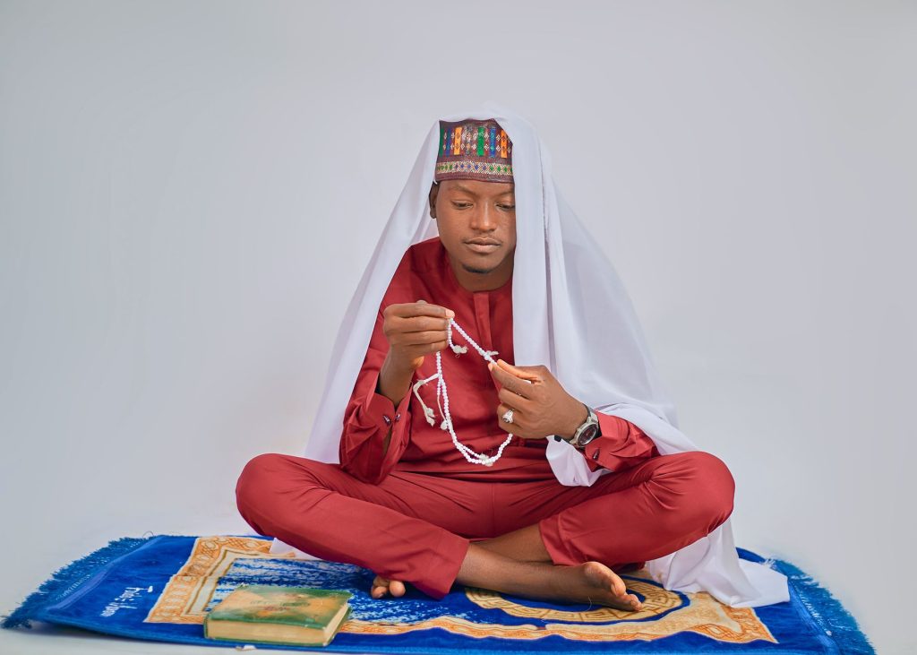 A young man in traditional attire sits cross-legged with prayer beads and a Quran, showing a moment of spiritual reflection.