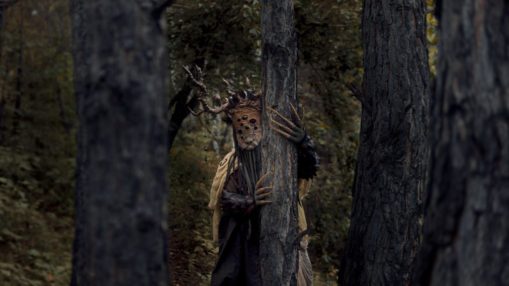 A mysterious and eerie forest scene with a costumed figure among the trees, evoking a sense of suspense. — Kantotan