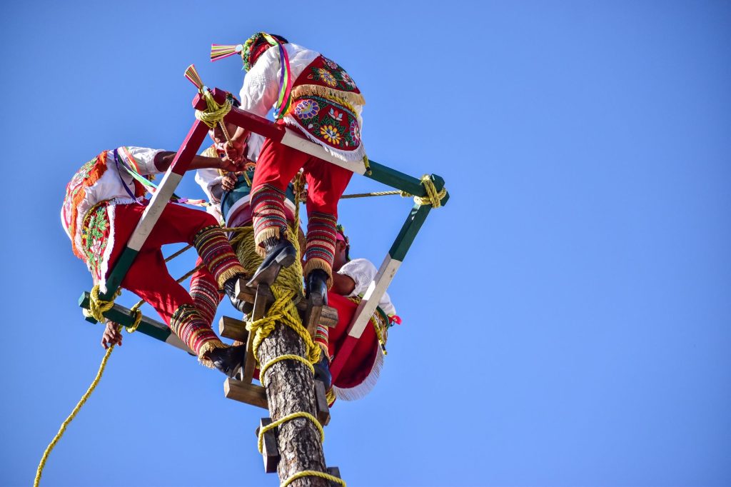 Performers in colorful traditional attire preparing for a ceremonial dance on a high wooden pole. — Kantotero