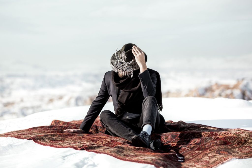 A stylish individual in a dramatic hat sits on a patterned rug amid Ortahisar's snowy landscape. — Kayatan.com