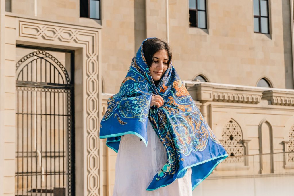 A woman wearing a colorful blue headscarf stands by an ornately detailed building facade on a sunny day. — Kayatan