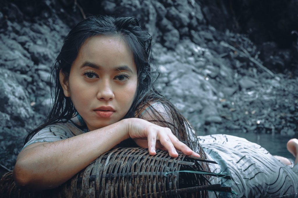 Attractive Asian female with wet hair looking at camera while lying in river with shabby wicker vase near shore in nature — M