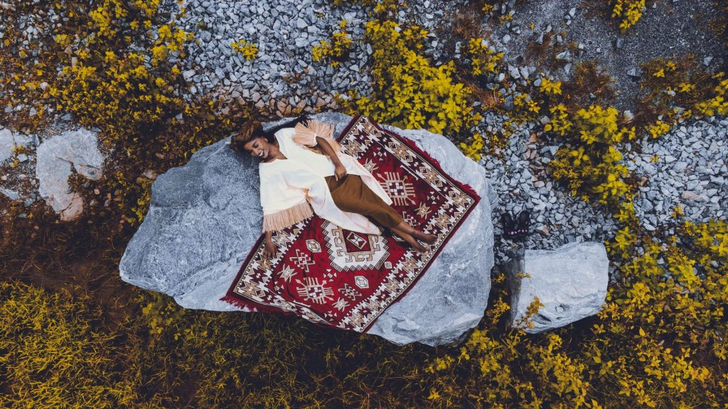 Aerial shot of a woman lying on a rug over a rock amidst nature with yellow foliage. — Mercury Rug