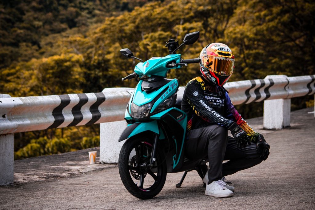 Motorcycle rider in vibrant gear poses with aqua motorbike on picturesque road. — Mio Sporty