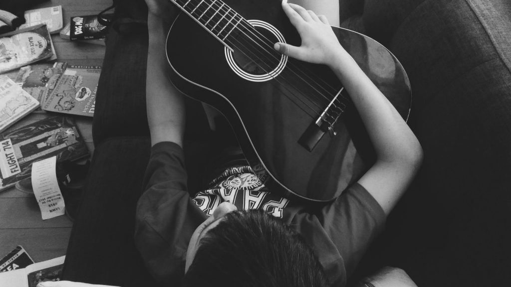 A young boy lying indoors on a couch playing an acoustic guitar with scattered books around. — Musicallydown