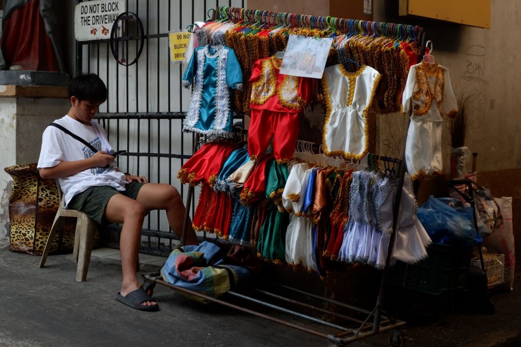 A street vendor sits beside a colorful display of traditional costumes, using a smartphone. — Nang At Ng