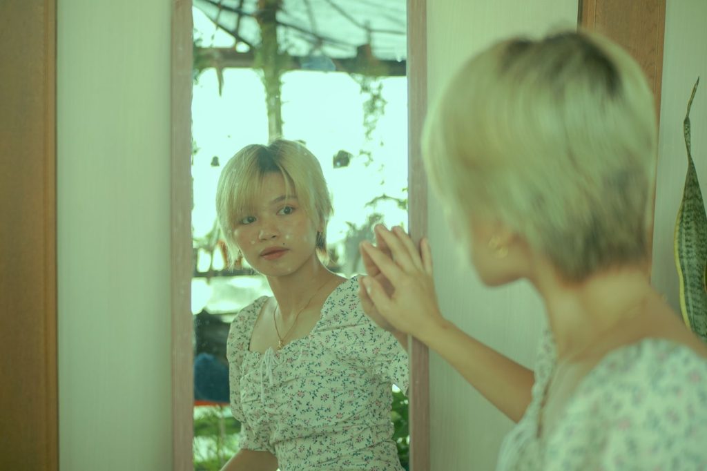 A woman with short blonde hair gazes at her reflection in a mirror inside a cozy room. — Paano