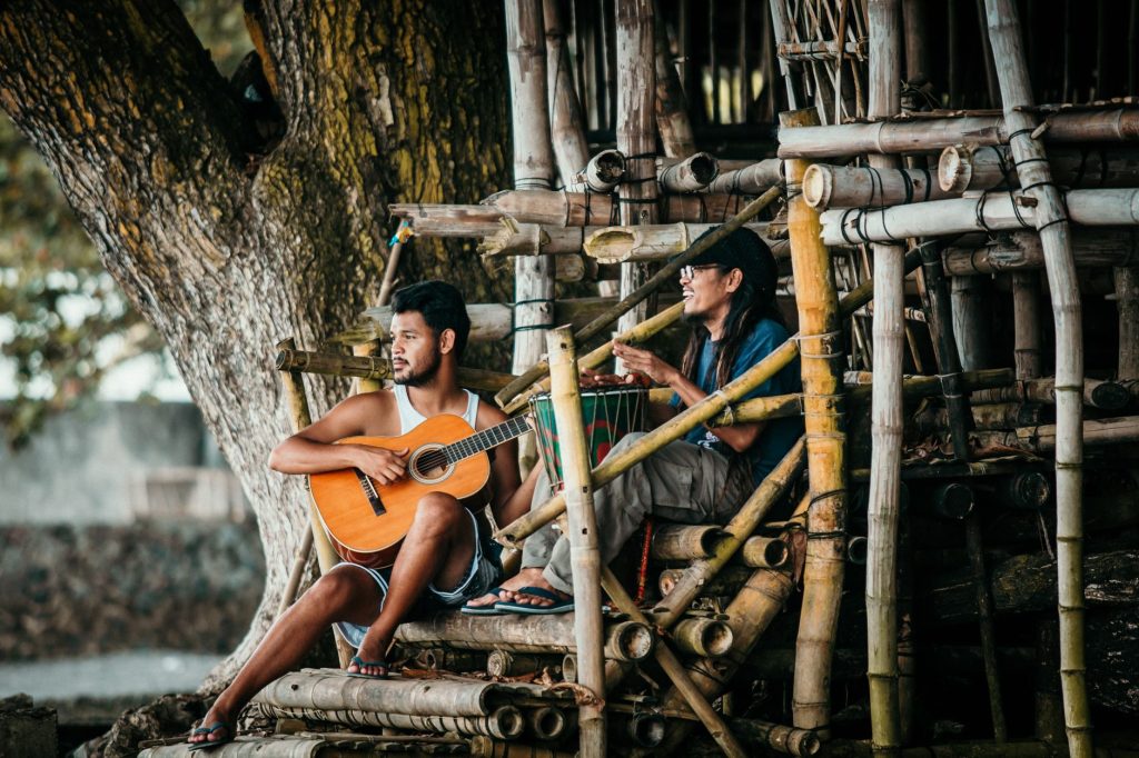 Young ethnic male guitarist playing classic guitar near cheerful partner with djembe on bamboo staircase near tree and lookin