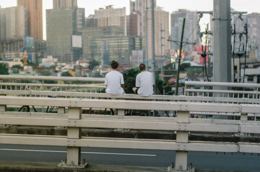 Two young adults sitting on a bridge, enjoying views of a bustling cityscape with skyscrapers. — Pasigpass