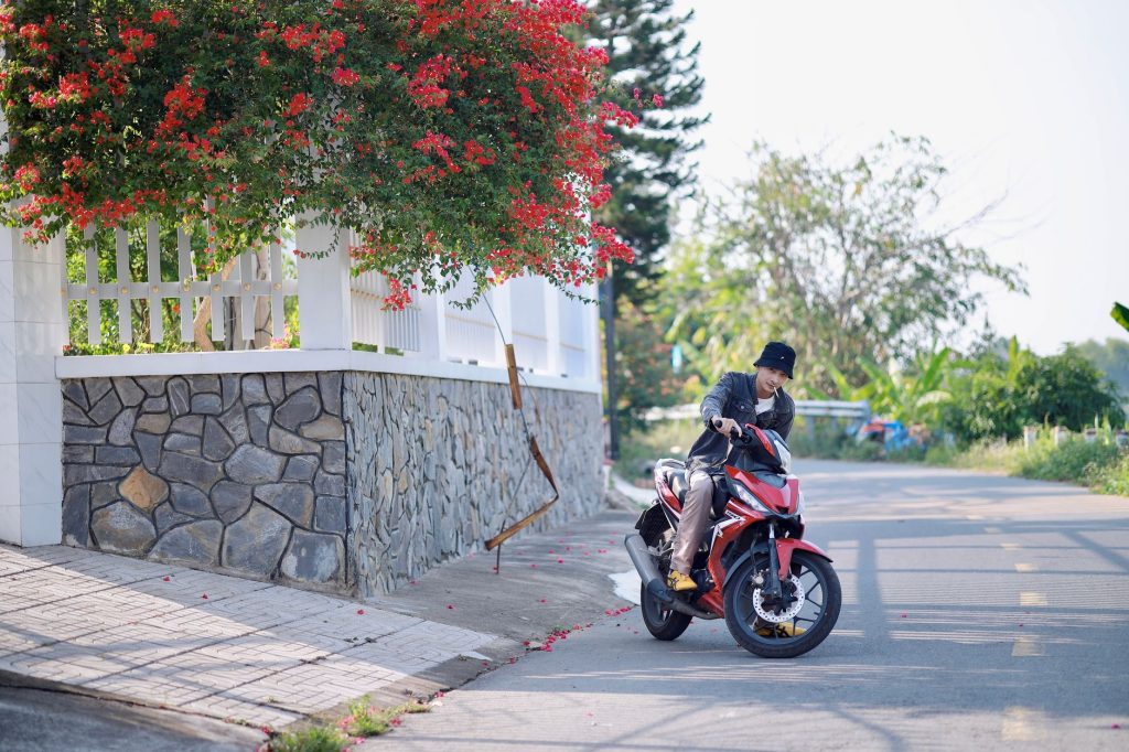 A man rides a motorcycle near a stone wall under blossoming trees on a sunny day. — Pcx 160
