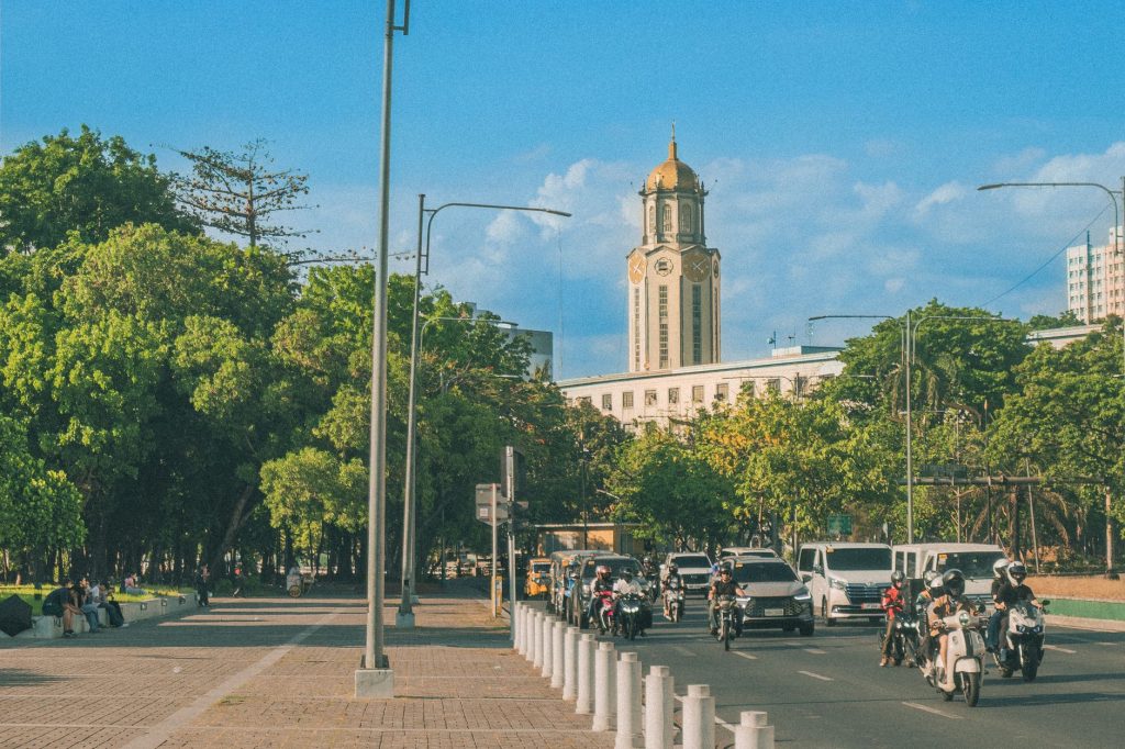 Bustling street in Manila with vehicles, lush trees, and the iconic clock tower under a clear blue sky. — Philgeps