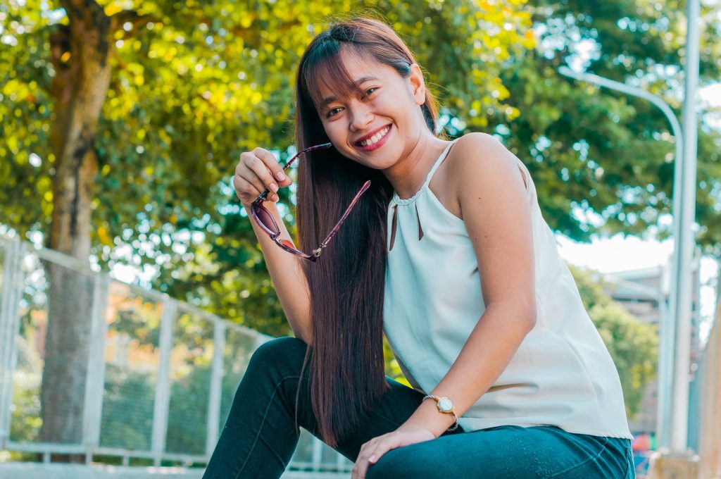 Delighted Asian female with long hair and sunglasses in hand smiling and looking at camera while sitting on street near green