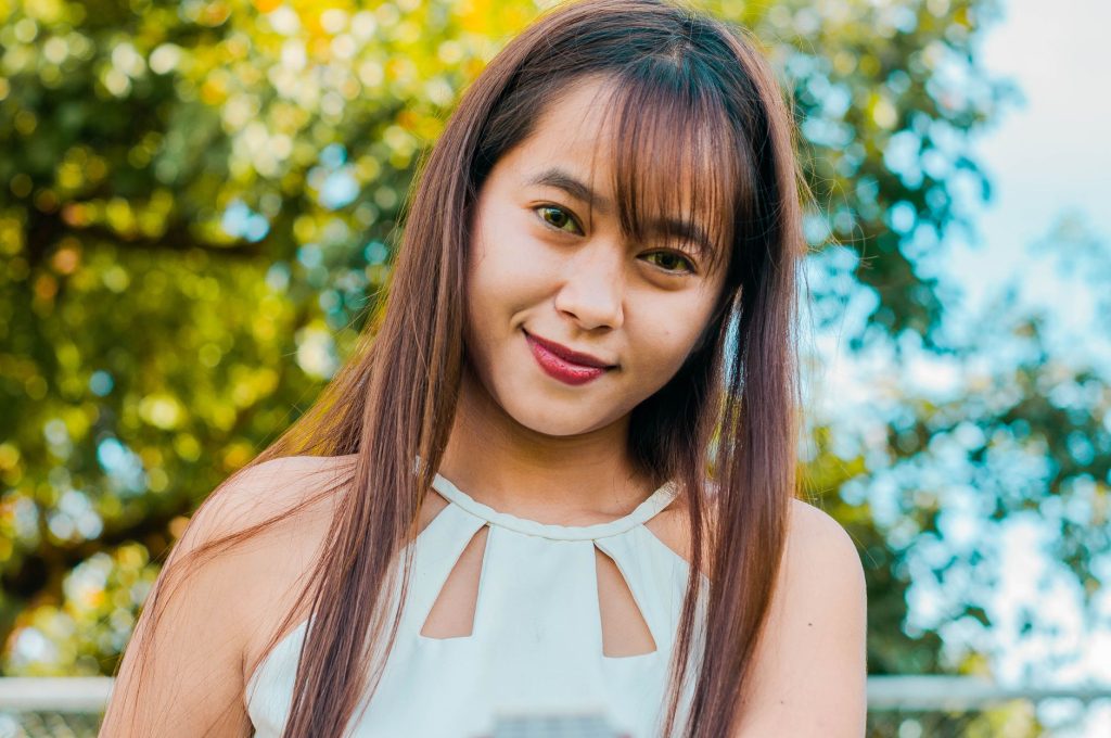 Positive Asian female with long hair looking at camera on street with tall lush trees on blurred background on summer day — P