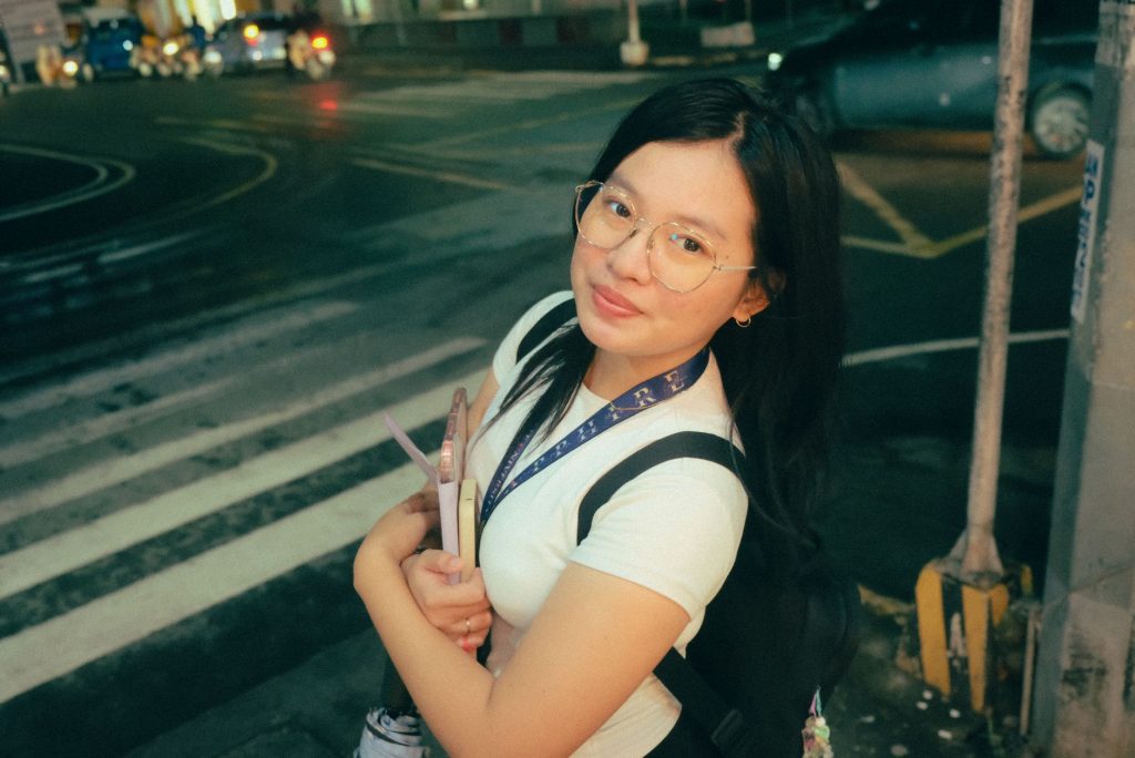A young woman with glasses smiles while standing at a crosswalk at night. — Pinay Pussy