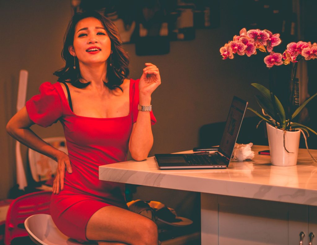 Chic woman in red dress smiling confidently at desk with laptop and orchids. — Pinayflex
