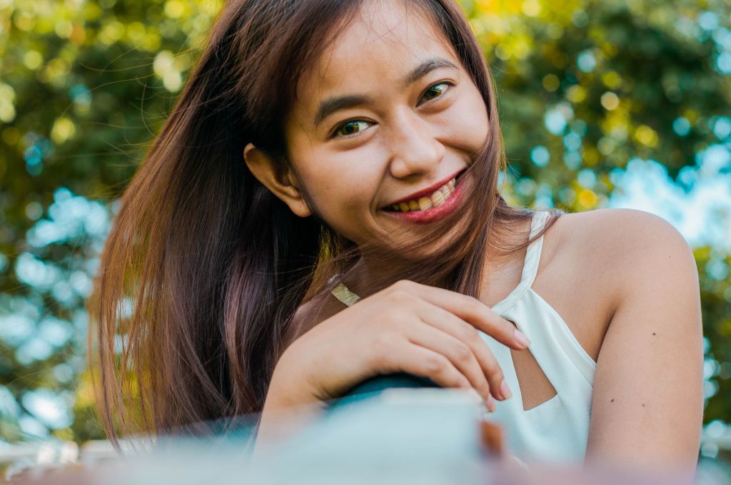 Positive Asian female with blurred ukulele in hand smiling and looking at camera on street with green trees on blurred backgr