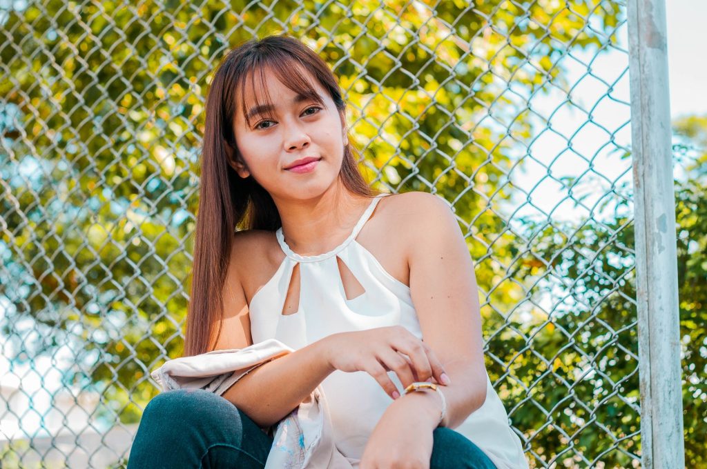 Content Asian female in stylish outfit looking at camera while sitting near metal grid on street with tree on sunny summer da