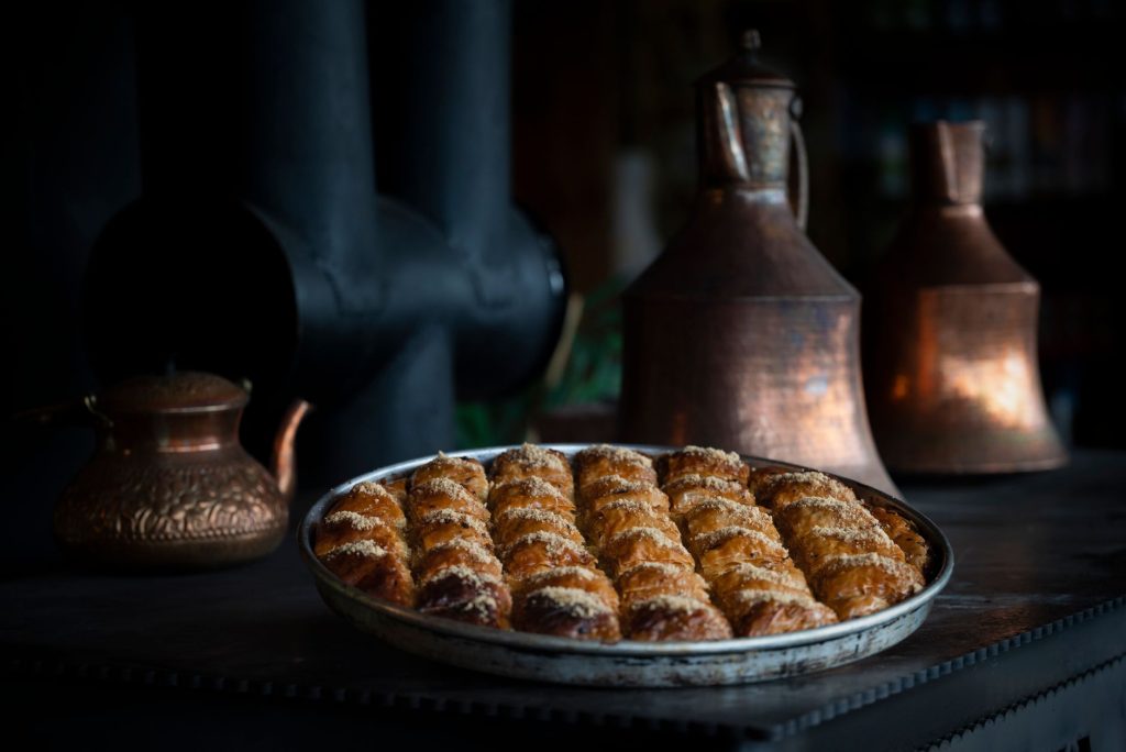A tray of freshly baked Turkish baklava displayed with antique copper pots in a dimly lit atmosphere. — Pisofi