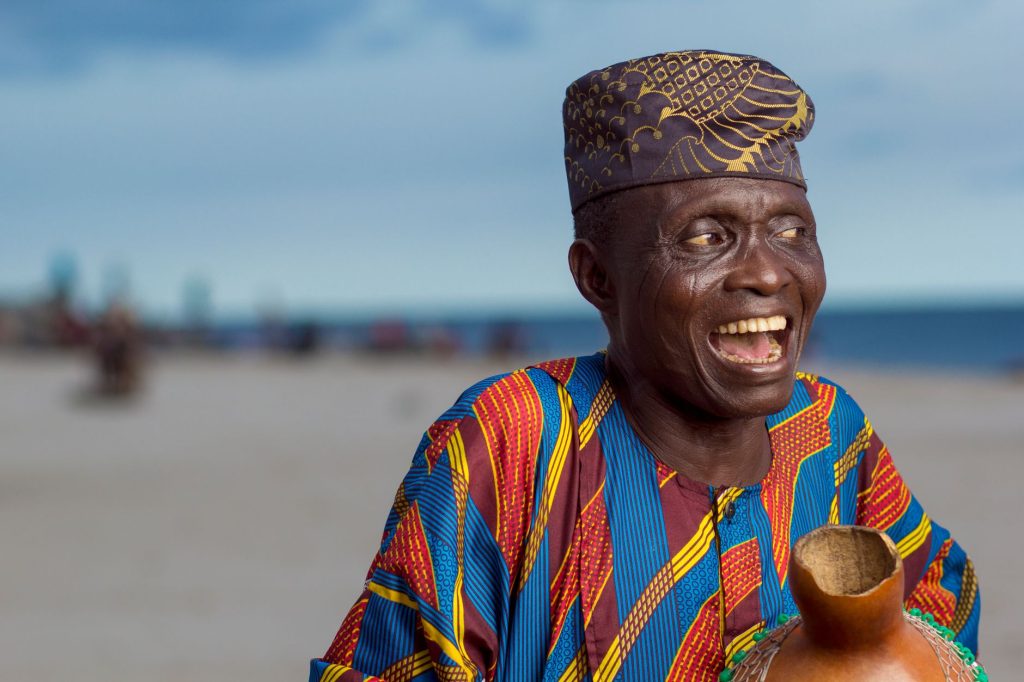 Elderly man joyfully smiling wearing Yoruba attire and cap on Lagos beach. — Pwetan