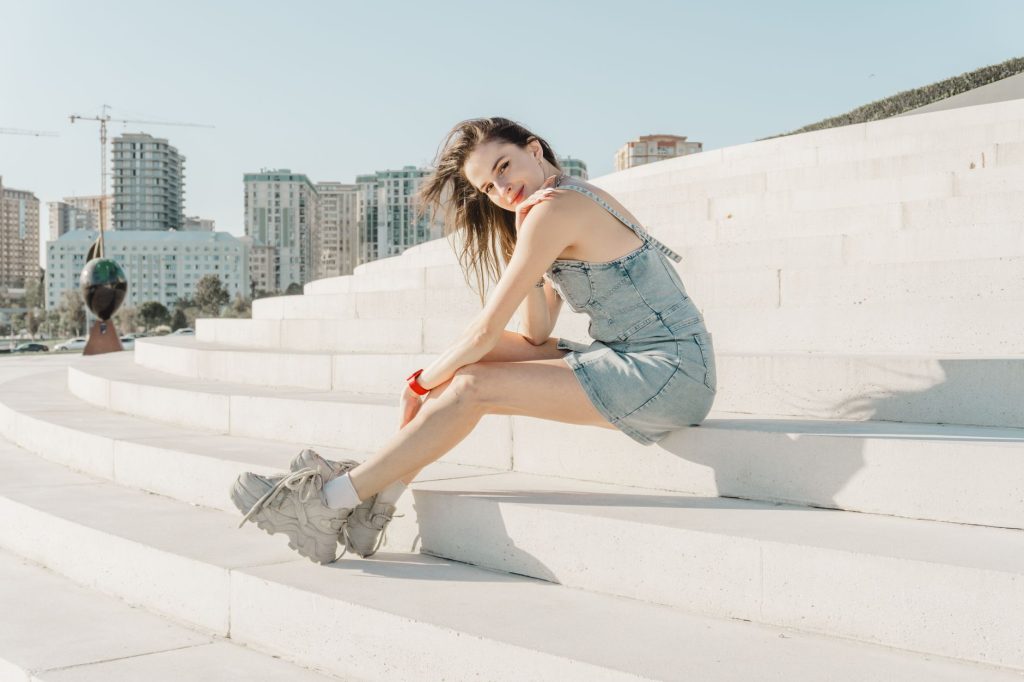 Stylish young woman sitting on white steps with cityscape in the background on a sunny day. — Rapbabe