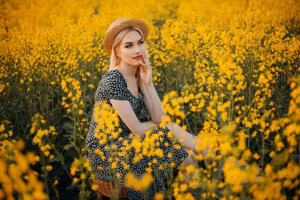 A woman in a floral dress and straw hat sitting in a field of yellow flowers, enjoying a peaceful moment. — Rapsababe