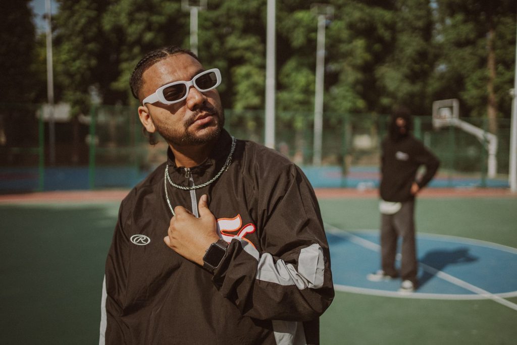 A fashionable man in sunglasses and streetwear poses confidently on a basketball court in New Delhi. — Rbreezy