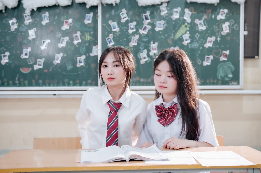 Two students in uniforms sitting at a desk with books in a classroom setting. — Secret Class