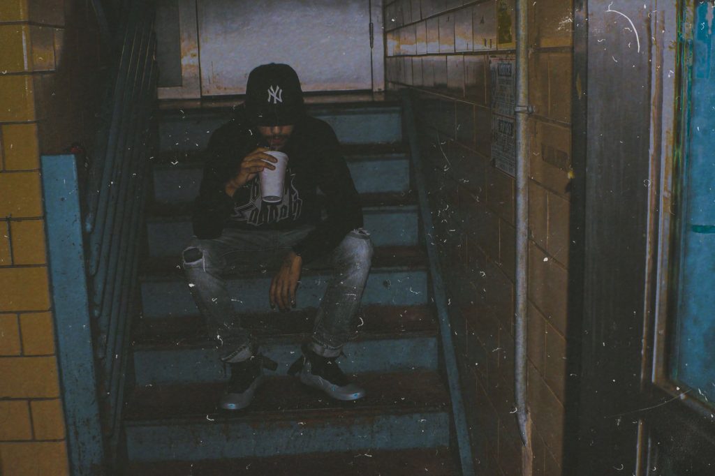 Man sitting on stairs in dimly lit urban setting, drinking from a paper cup. — Tmtplay