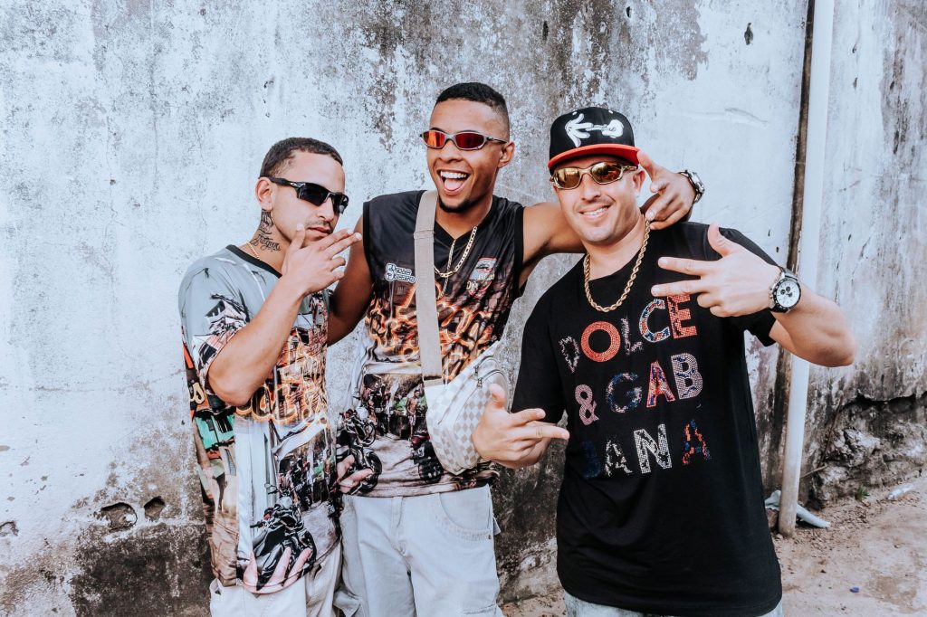 Three urban musicians posing energetically against a rustic wall, showcasing streetwear fashion. — Ujiz