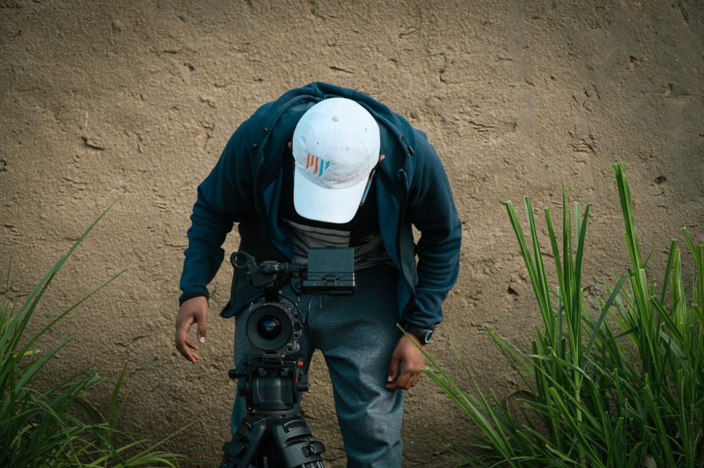 A man adjusts a camera setup outdoors near a grassy wall. — Ujizz