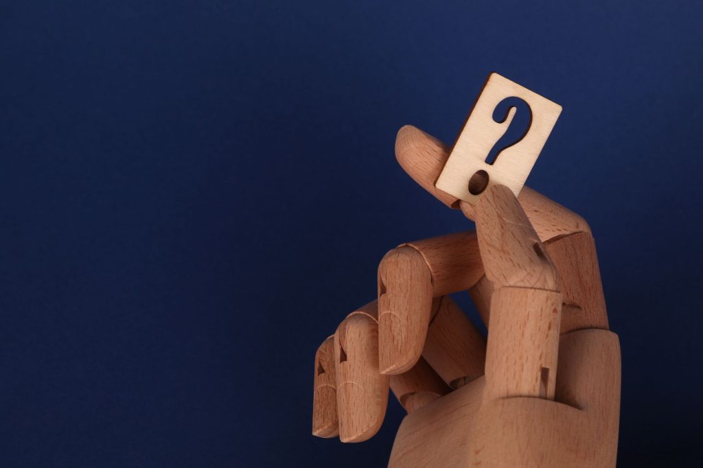Wooden mannequin hand holding a block with a question mark against a blue background. — Which Of The Following
