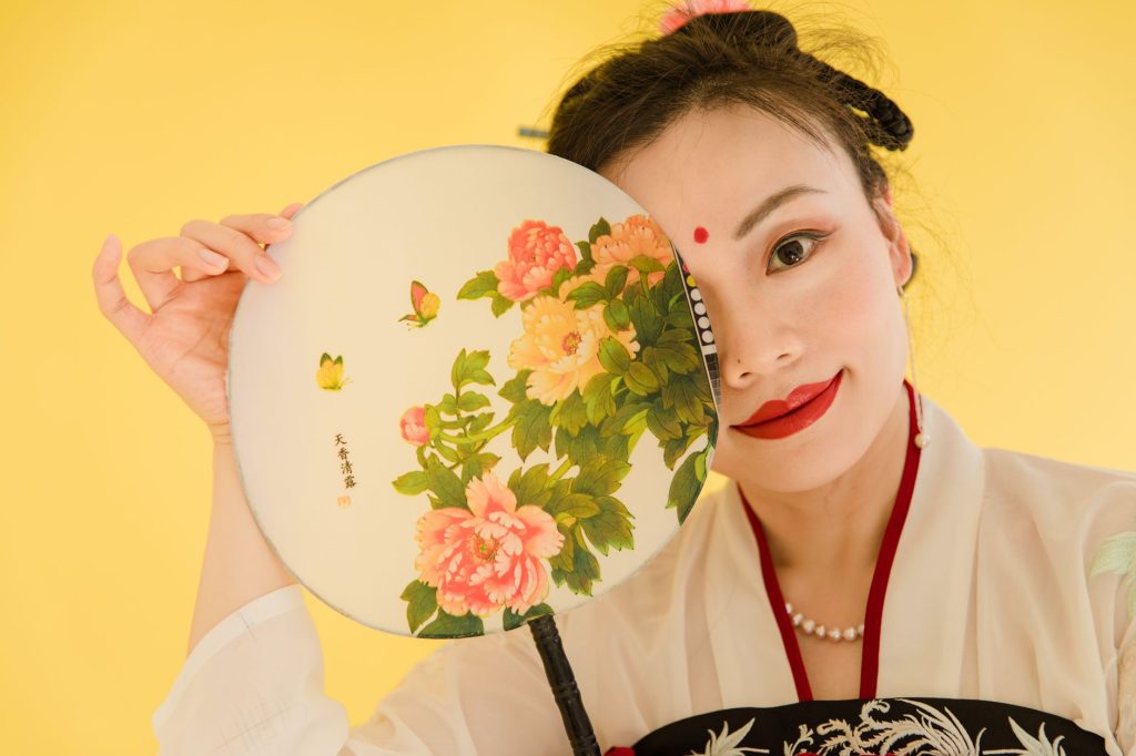 Portrait of an Asian woman in traditional attire holding a floral hand fan against a yellow background. — Yujiz